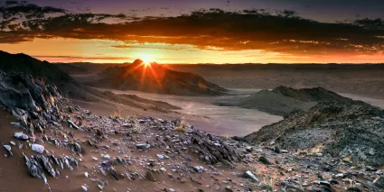 A vivid sunset viewed from the summit of White Mountain, looking west across the infinite orange dunes of the Namib Desert toward a horizon of fire and dust.