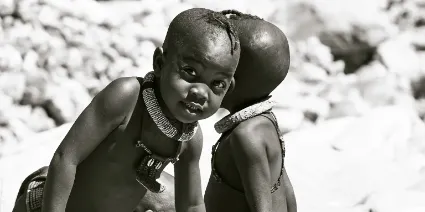 A monochrome study of two Himba boys at a natural spring rock pool, documenting the carefree and tactile connection to the land's essential resources.