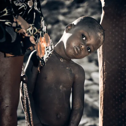 An inquisitive Himba child peeking from behind his mother, a candid study of childhood curiosity in the Kunene region.