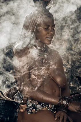 A Himba woman partially cloaked in woodsmoke while preparing a meal, a narrative of daily life and the ancient rhythms of the domestic hearth.