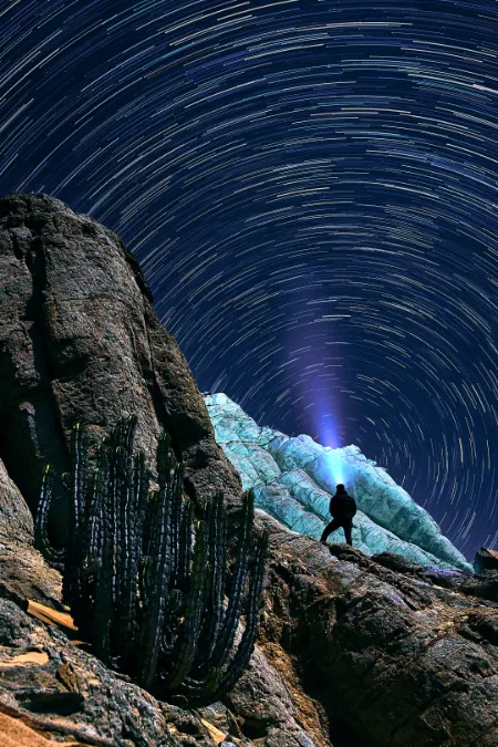 Star trails above Rössing Mountain near the coast; a long-exposure study featuring a lone figure illuminating the night sky.