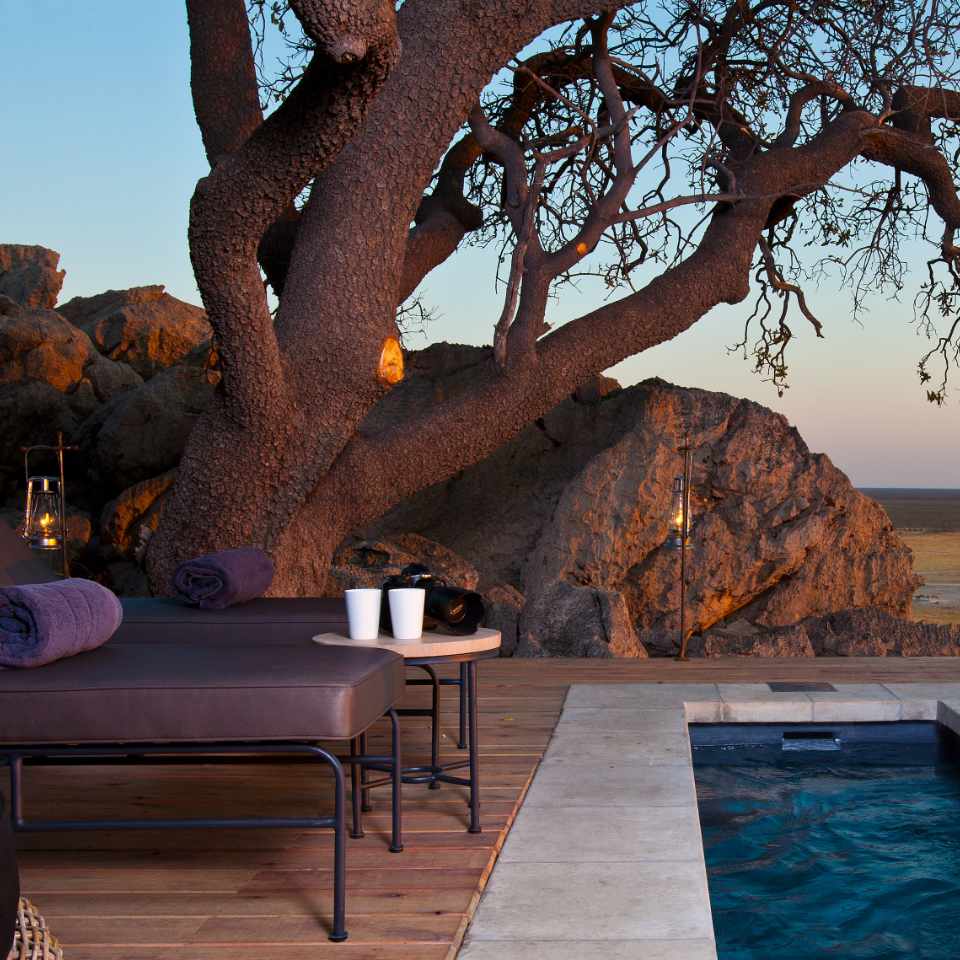 Twilight serenity at Dolomite Camp's luxury chalets, featuring a majestic tree, ambient lanterns, and an inviting pool with panoramic views of Etosha.