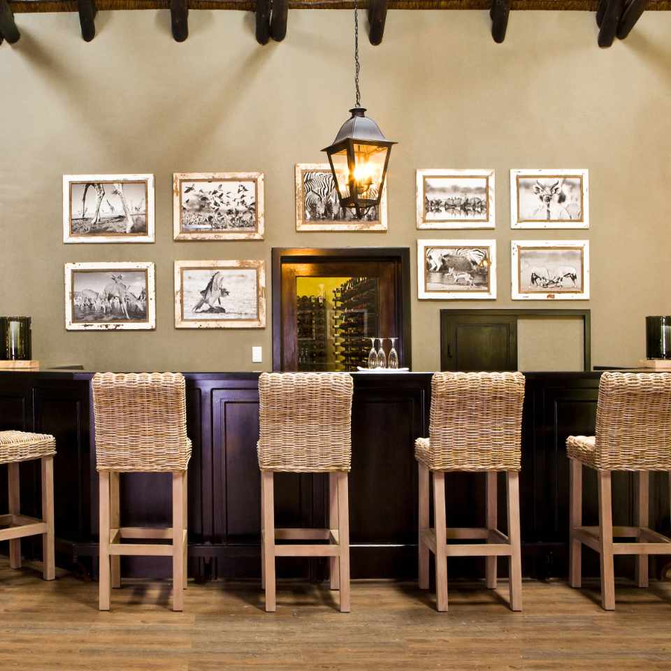 Elegant bar area at Mushara Lodge with rustic charm, featuring high wooden chairs, framed black and white photographs, and a warm lantern-style pendant light.