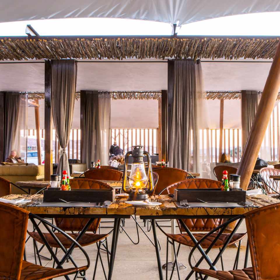 Rustic elegance in the dining area of Dead Valley Lodge, featuring thatched roofing, lantern-lit wooden tables, and long curtains for an intimate dining experience.