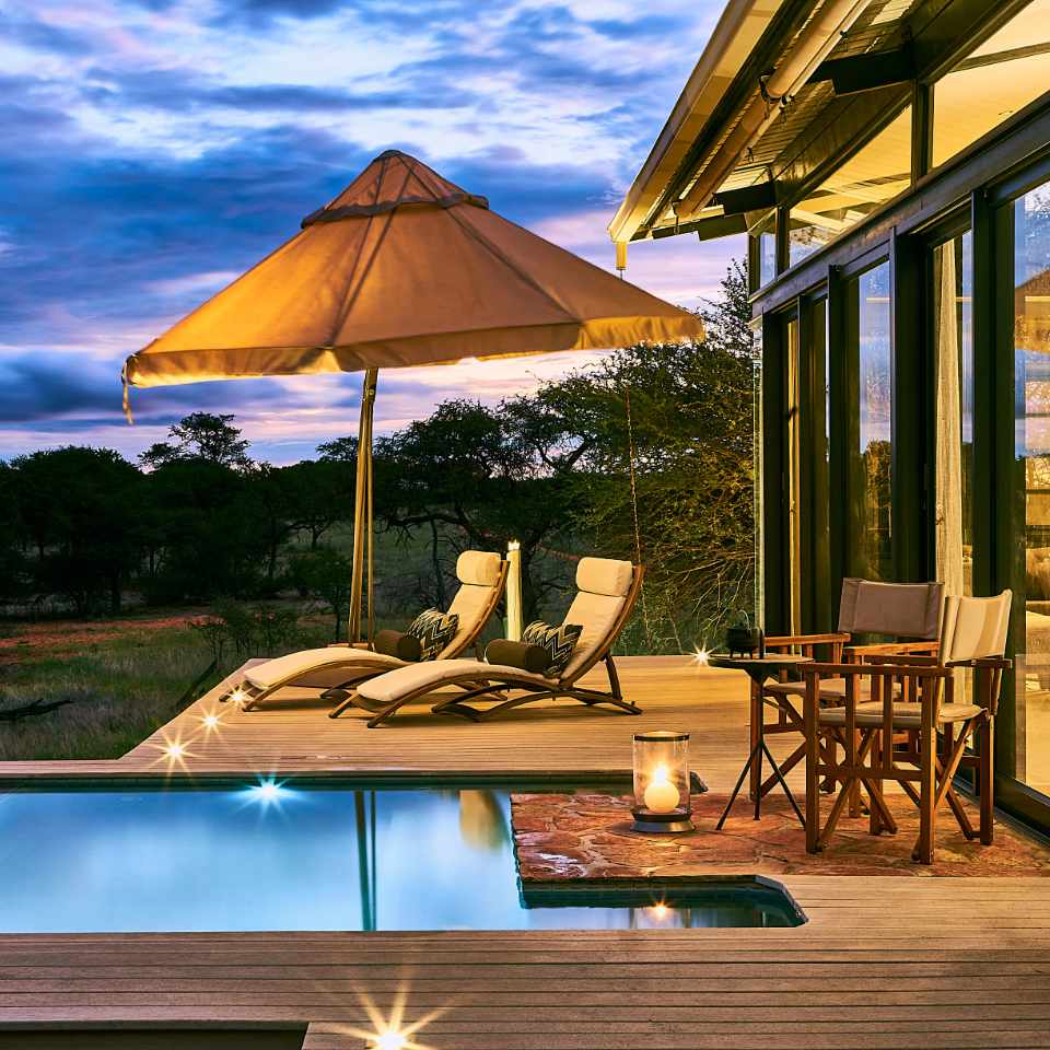 Opulent outdoor relaxation at Ozimbandunda Lodge's luxurious suite, featuring a tranquil pool, elegant lounge chairs, and breathtaking sunset views in central Namibia.