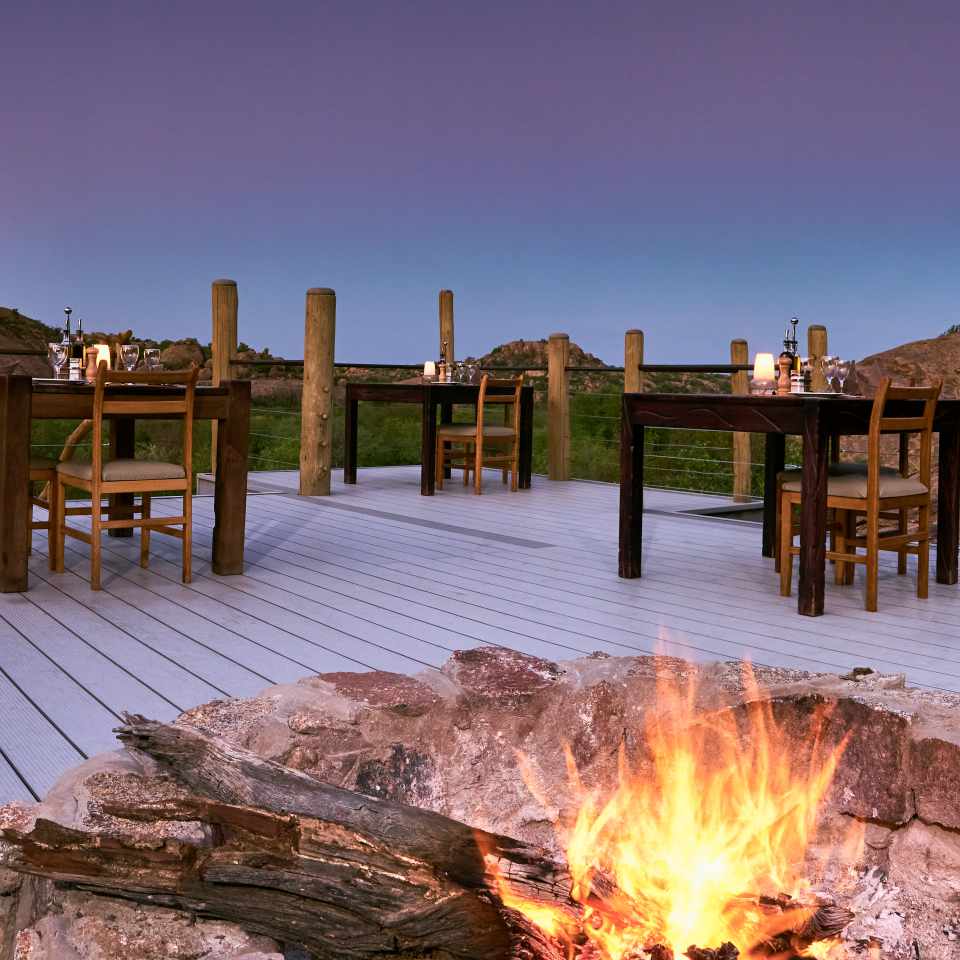 Inviting outdoor dining setup at Erongo Wilderness Lodge with wooden tables, a cozy fire, and a stunning backdrop of Erongo Mountains at twilight.