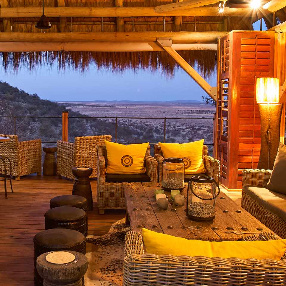 Inviting outdoor bar area at Dolomite Camp, Etosha National Park, featuring wicker chairs with yellow cushions under a thatched roof, offering a panoramic view of the Namibian wilderness at twilight.