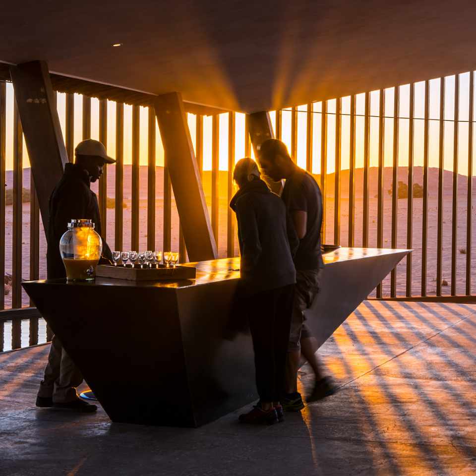 Warm sunset hues silhouette guests checking in at Dead Valley Lodge, with a modern counter and panoramic desert views through open walls.