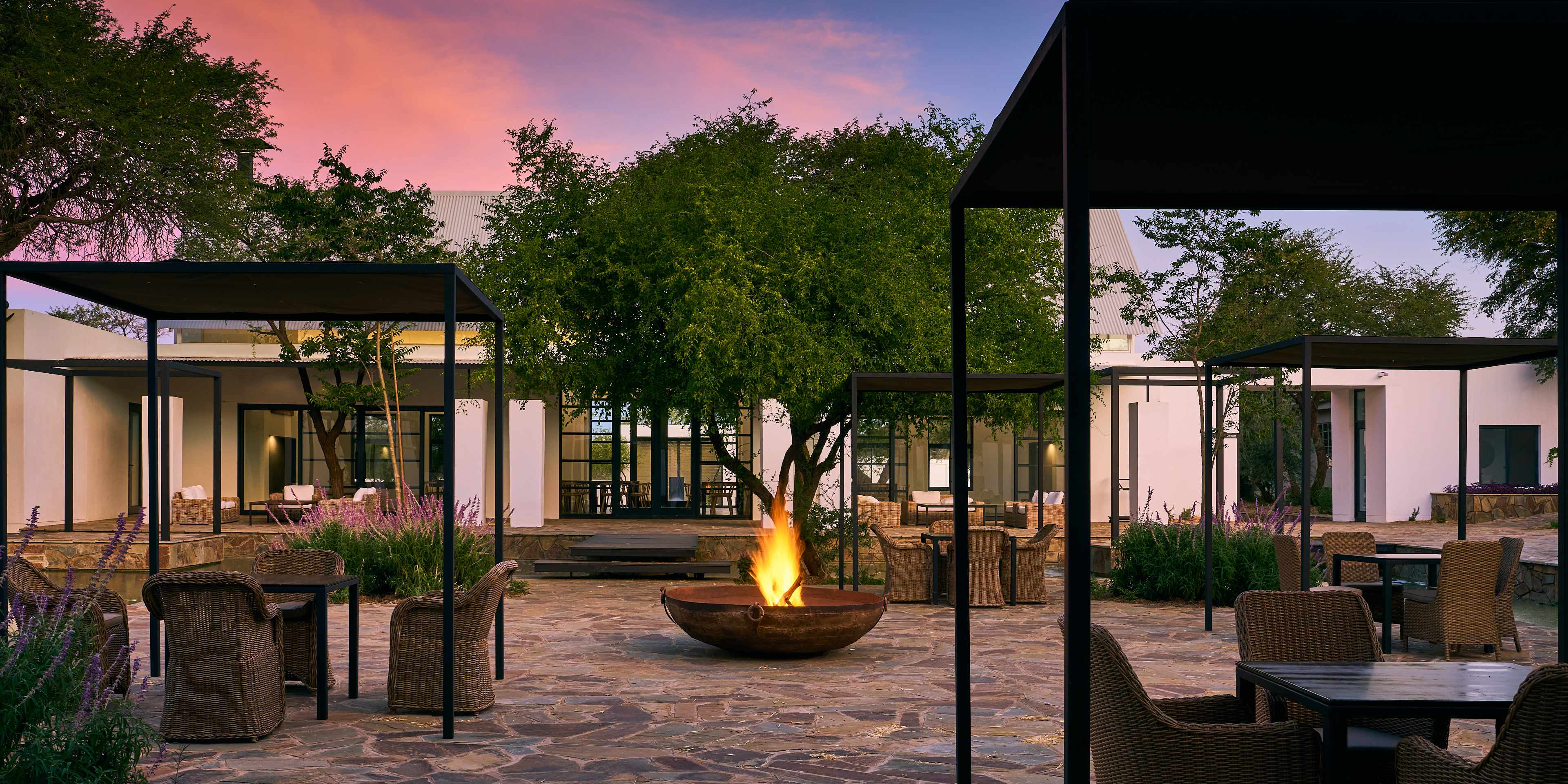 Stunning evening view of Sandwerf Lodge, a passion project by Namibia's leading architect, featuring modern design with large glass windows and a cozy fire pit amidst lush greenery.