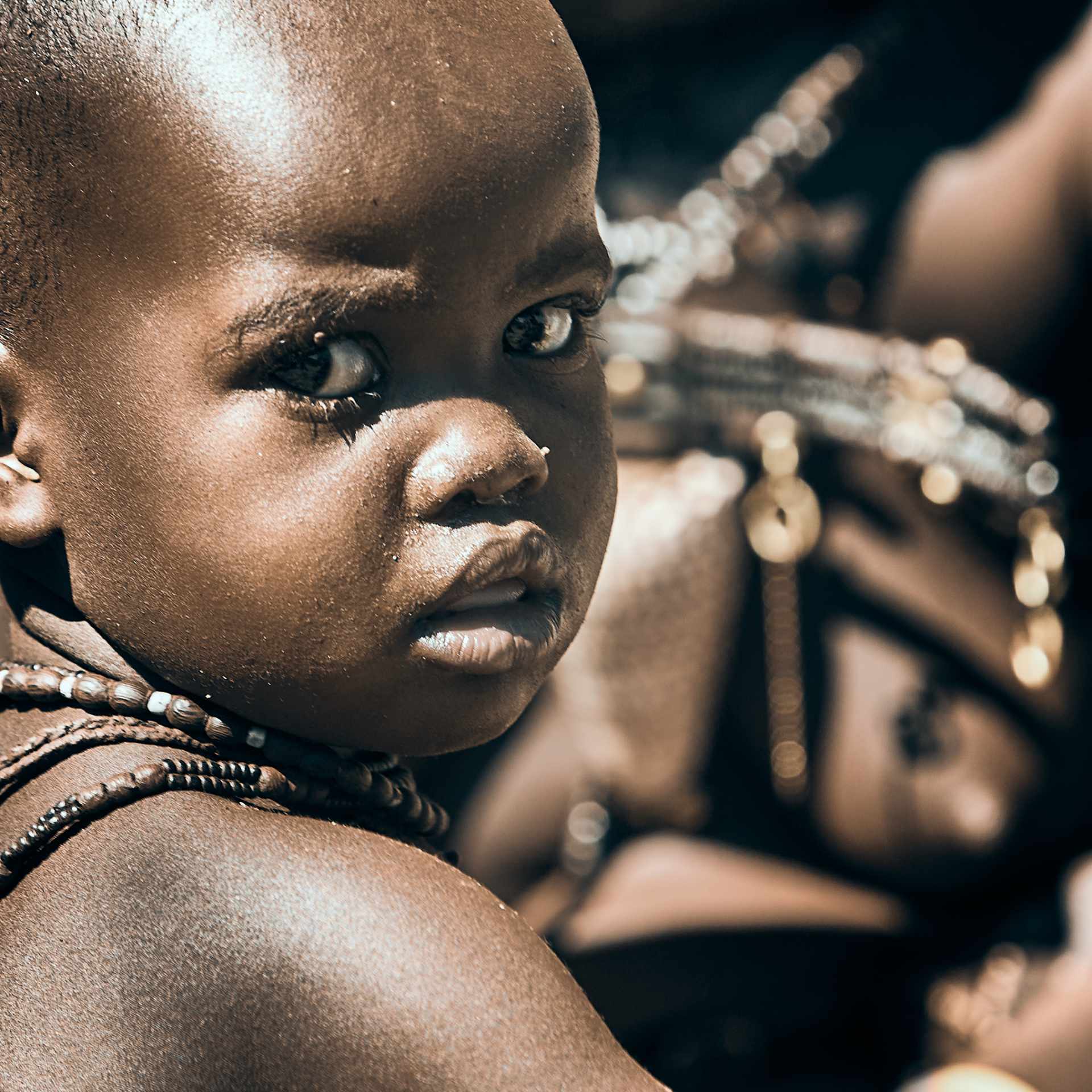 Young Himba Life: A candid moment capturing a young Himba boy adorned with traditional jewelry, his skin glistening in the Namibian sun, reflecting the vibrant culture and heritage of his people.