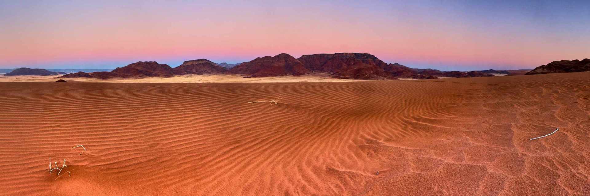 Vibrant blue hour over the dunes of Sossusvlei, with a mountain range breaking the horizon under a bluish-pink sky, and sand ripples leading the viewer's eye into the distance.